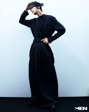 Male fashion model poses in a full-length black garment, complemented by a matching mask and belt, against a stark white backdrop.