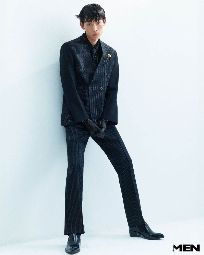 A slim adult male poses in a dark pinstripe suit in front of a light blue wall, wearing leather gloves and patent leather dress shoes in studio.