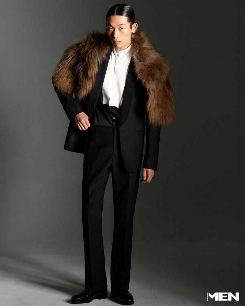 Man in black suit and fur stole stands in studio. Dark hair slicked back, looking at camera. Fashion, style, and formalwear.