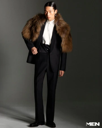 Man in black suit and fur stole stands in studio. Dark hair slicked back, looking at camera. Fashion, style, and formalwear.