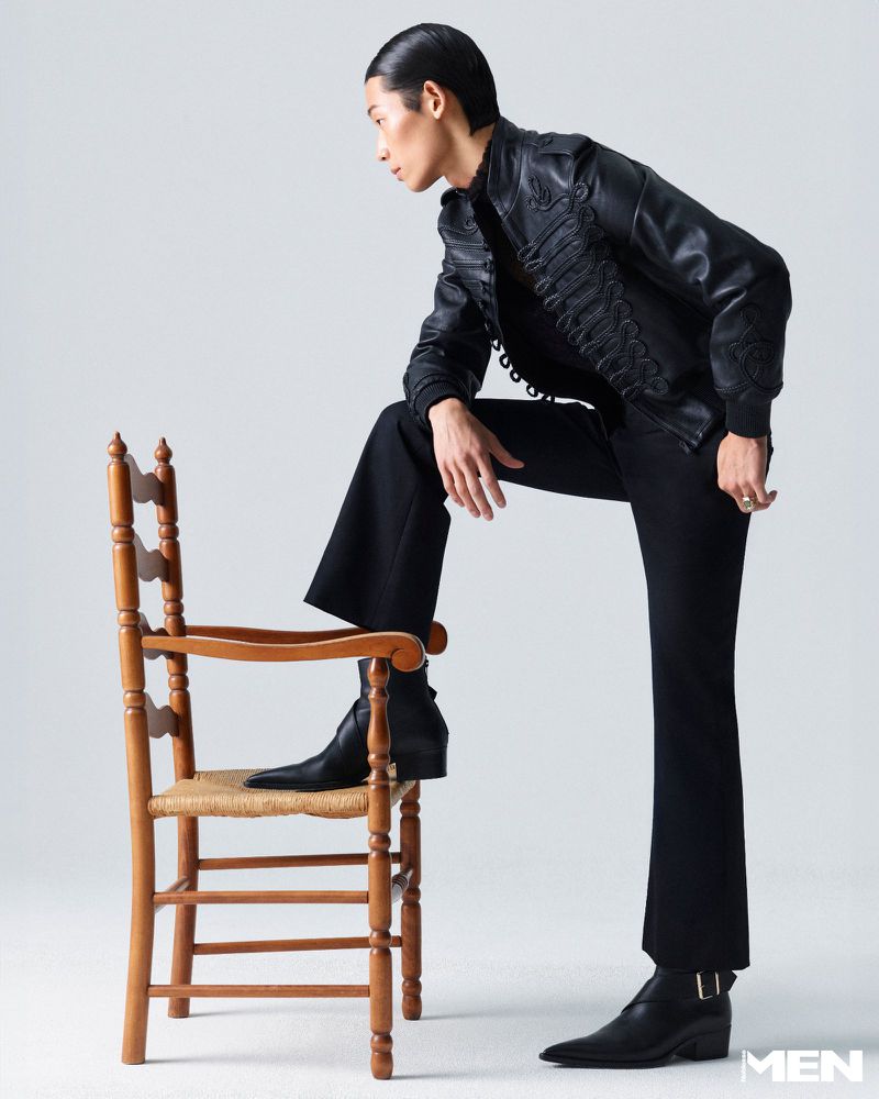 Model Min Seok Choi poses in an ornate black leather jacket and boots with one foot resting on a wooden chair against a studio backdrop.