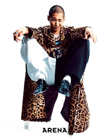 Stylish adult male poses seated, wearing a leopard print coat, patchwork sweatpants, and shiny black dress shoes in a studio.
