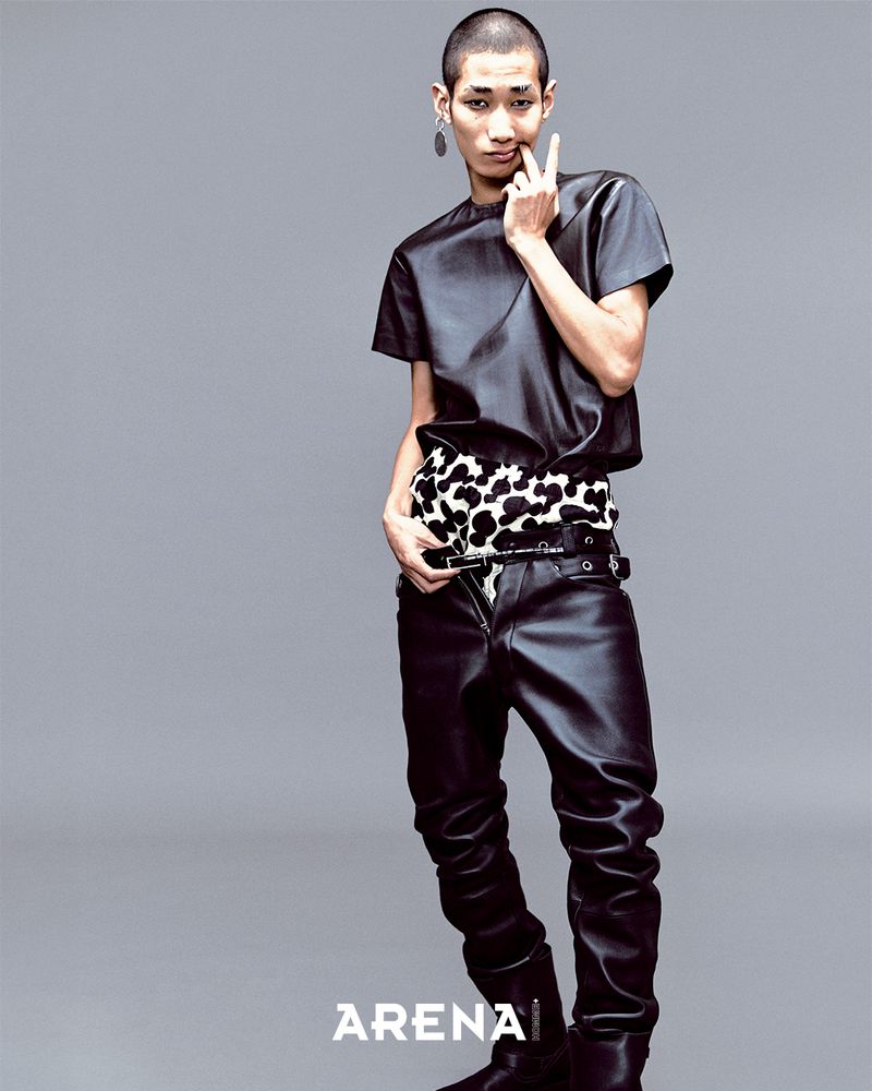 A person in a black leather-like outfit poses in a studio with a gray background. They are wearing an animal print accessory.