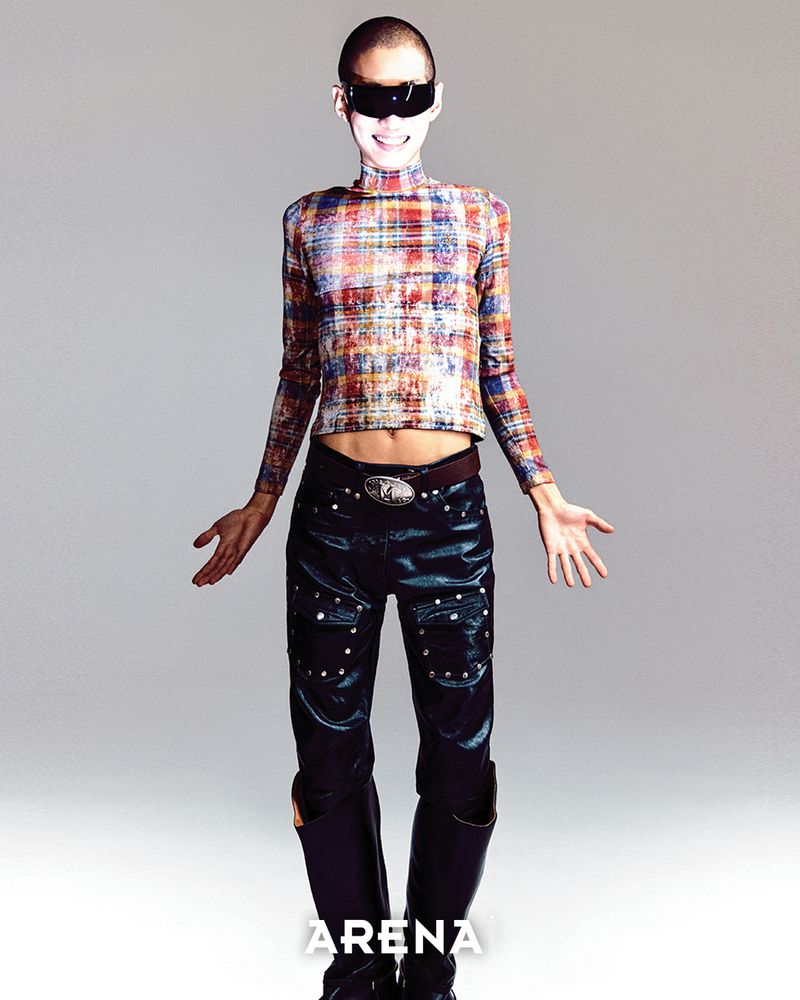 A slender person with a shaved head, stylish sunglasses, and patterned top poses in a studio setting with dark trousers and boots.