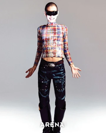 A slender person with a shaved head, stylish sunglasses, and patterned top poses in a studio setting with dark trousers and boots.
