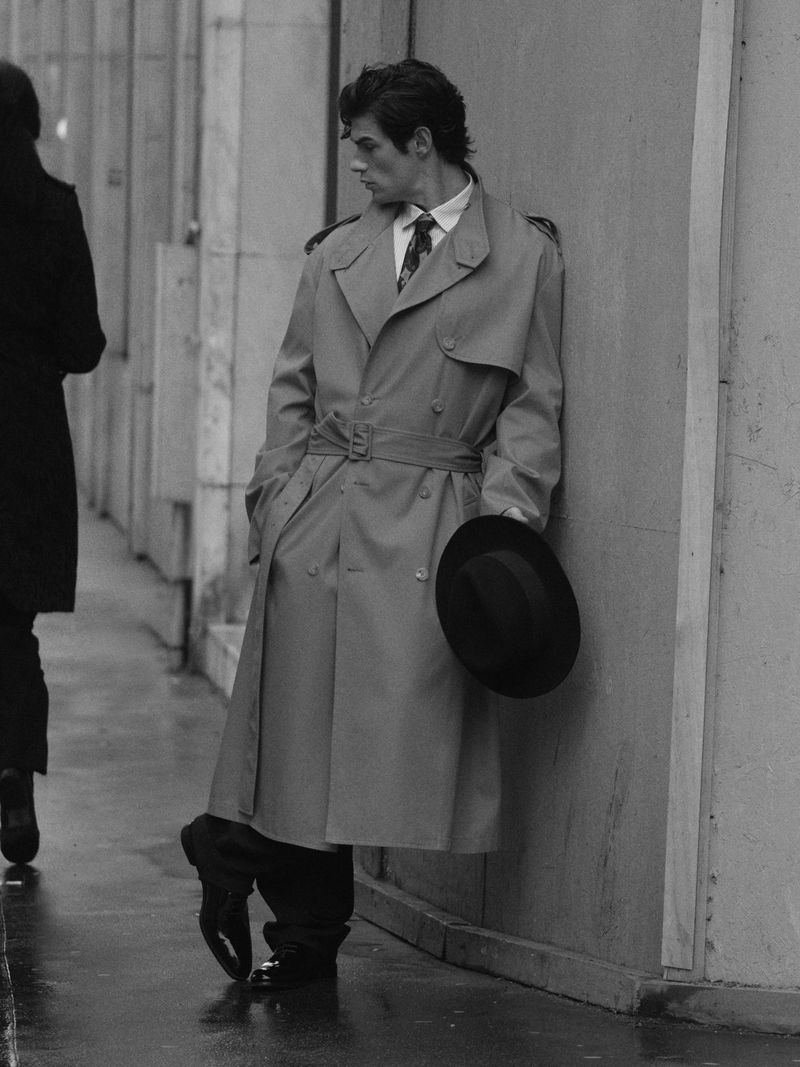 Monochrome portrait of a stylish adult man wearing a trench coat, leaning against a wall and holding a hat on a wet urban street.