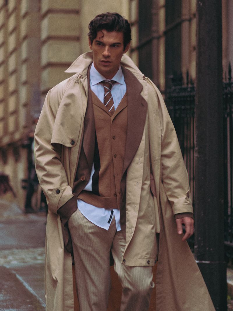 Male model in tan trench coat and brown suit walking outdoors against a building. Fashionable urban style with a sophisticated look.