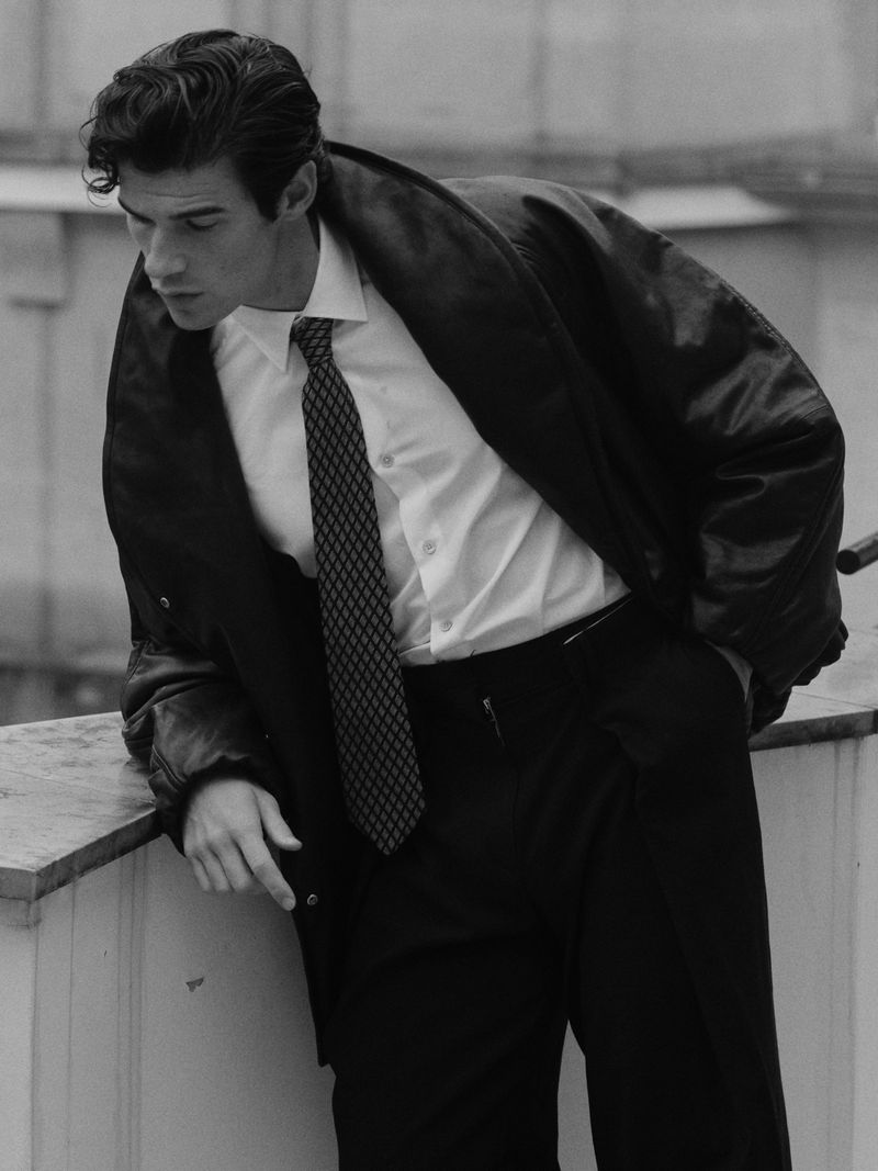 Black and white portrait of an adult man wearing a dress shirt, necktie, slacks and jacket, leaning against a ledge in an urban environment.