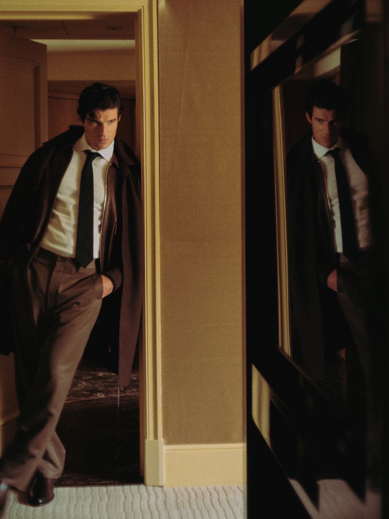 A stylish man in a suit and trench coat stands in a doorway with a neutral wall, softly lit in an indoor setting, reflected in a dark mirror.