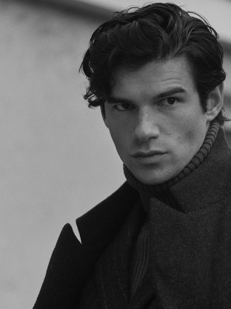 Portrait of a brunette adult male in a turtleneck sweater and coat, looking directly at the camera in a monochrome photograph.