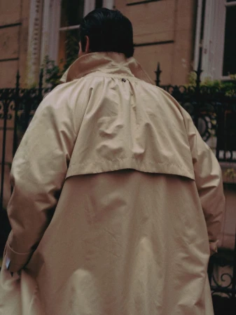 Person with dark hair in tan trench coat walking on sidewalk in front of building with arched window and garden plants.