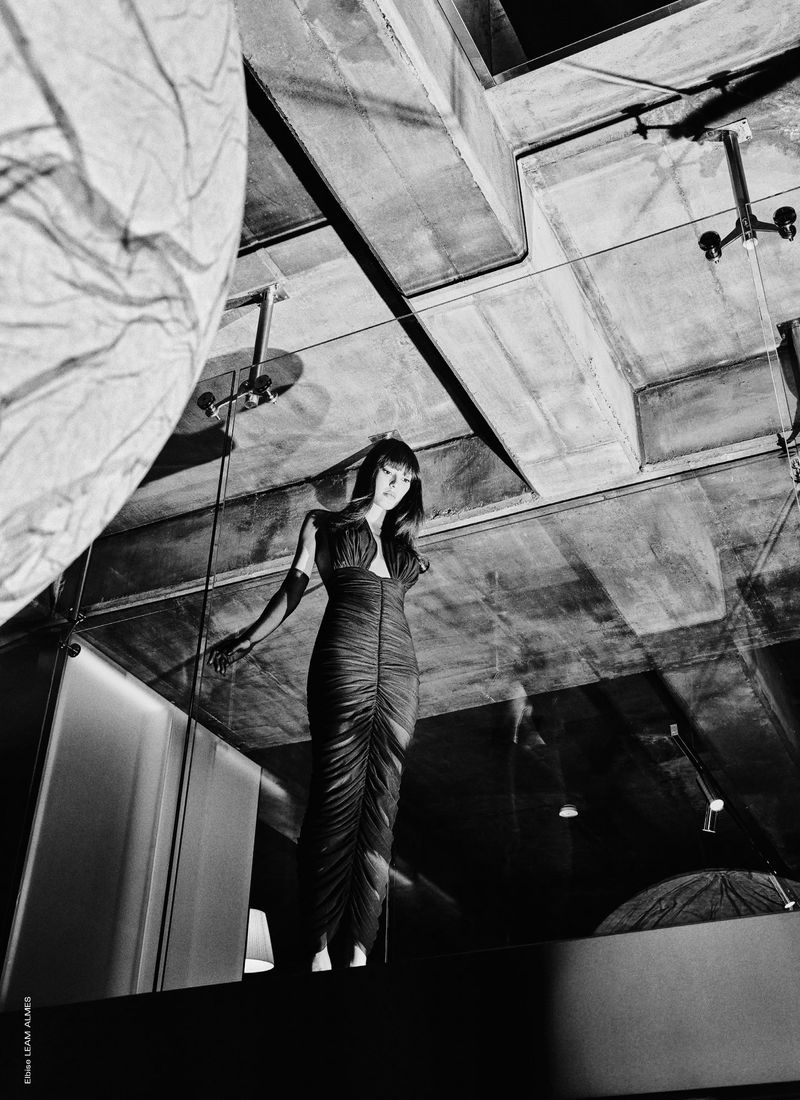 A long-shot black and white photograph of a woman in a long dress, posing in an indoor environment with abstract interior design.