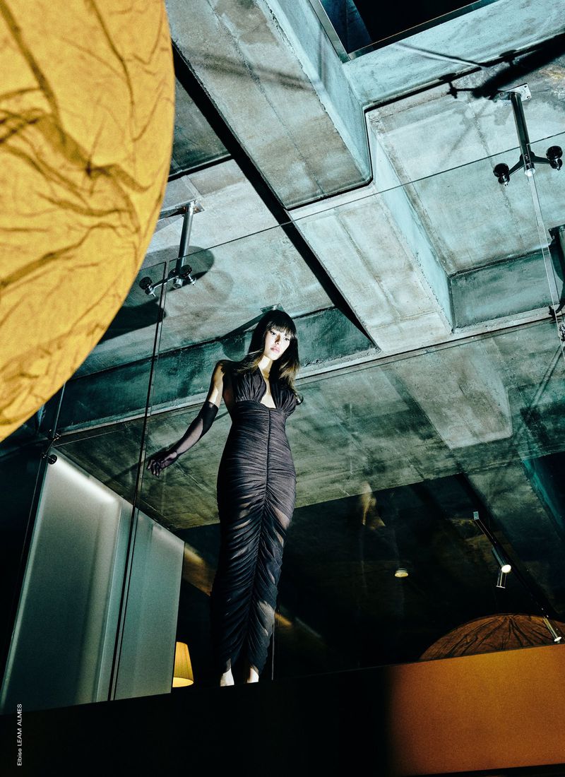 Elegant woman in a black dress stands in a modern, dimly lit interior with glass walls, concrete ceilings, and unique lighting fixtures.