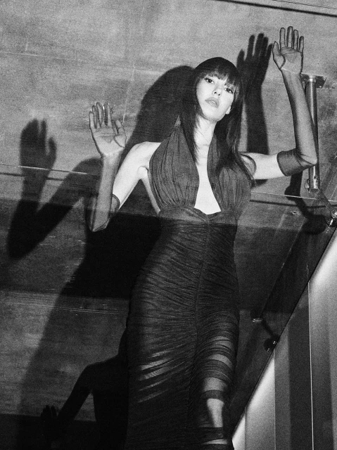 Monochrome portrait of a woman in an evening gown with hands raised against a textured wall, casting dramatic shadows in a low-light environment.