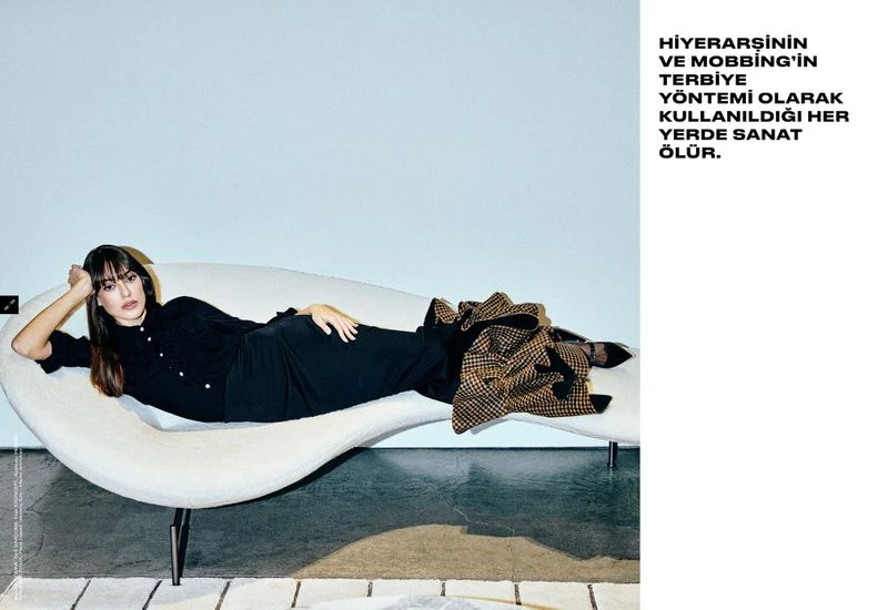 Brunette woman lies on white couch in black dress with tan patterned coat, indoors against a light blue wall.