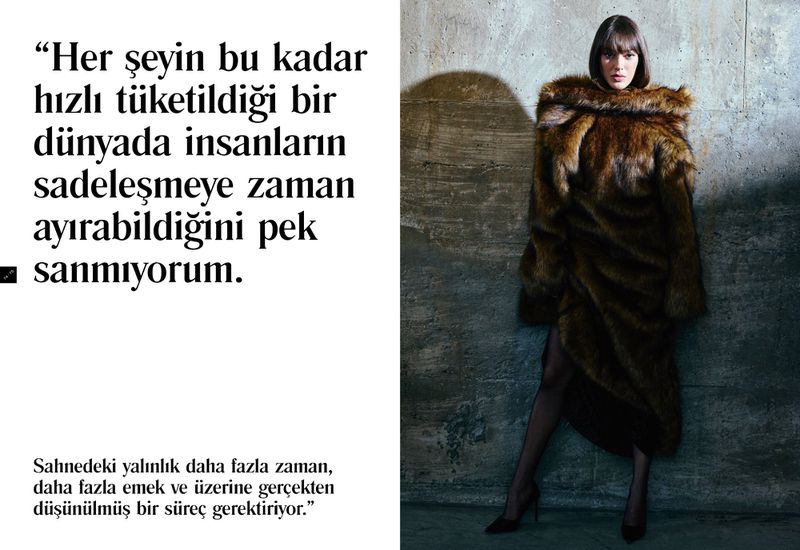Elegant woman in a long fur coat posing indoors against a textured wall, showcasing fashion and style, with soft studio lighting.