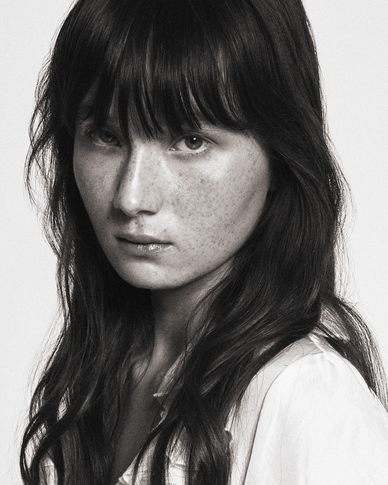 Striking black and white close-up portrait of a woman with freckles, dark hair, and a white shirt against a plain background.