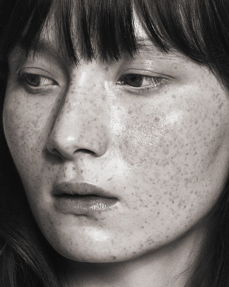 Close-up portrait of an adult female with fair skin, dark hair, and freckles, looking down with a neutral expression in soft lighting.