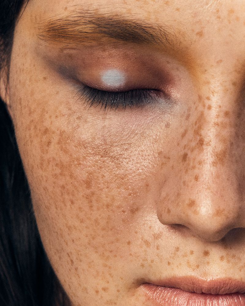 Close-up on an adult woman's face featuring an eye with white makeup, freckled skin, eyelashes, eyebrow, nose, and mouth.