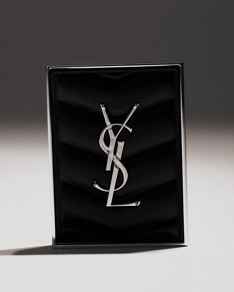 Silver YSL logo on a black leather textured wallet or card holder, against a gradient gray background in a studio product shot.