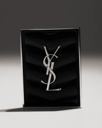 YSL black quilted makeup compact with a silver interlocking monogram logo standing on a neutral grey studio background.