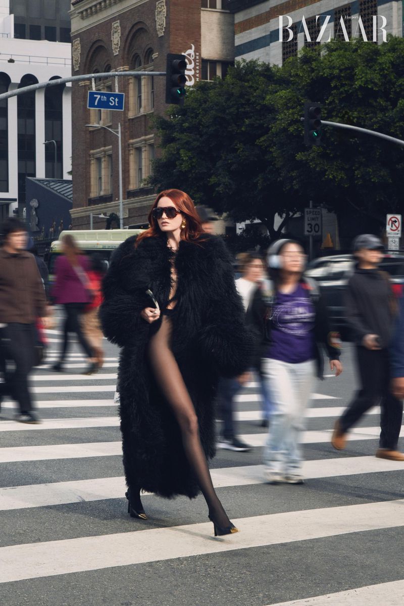Woman in fur coat walks along crosswalk in city street with blurred pedestrians; Harper's Bazaar logo visible in background.