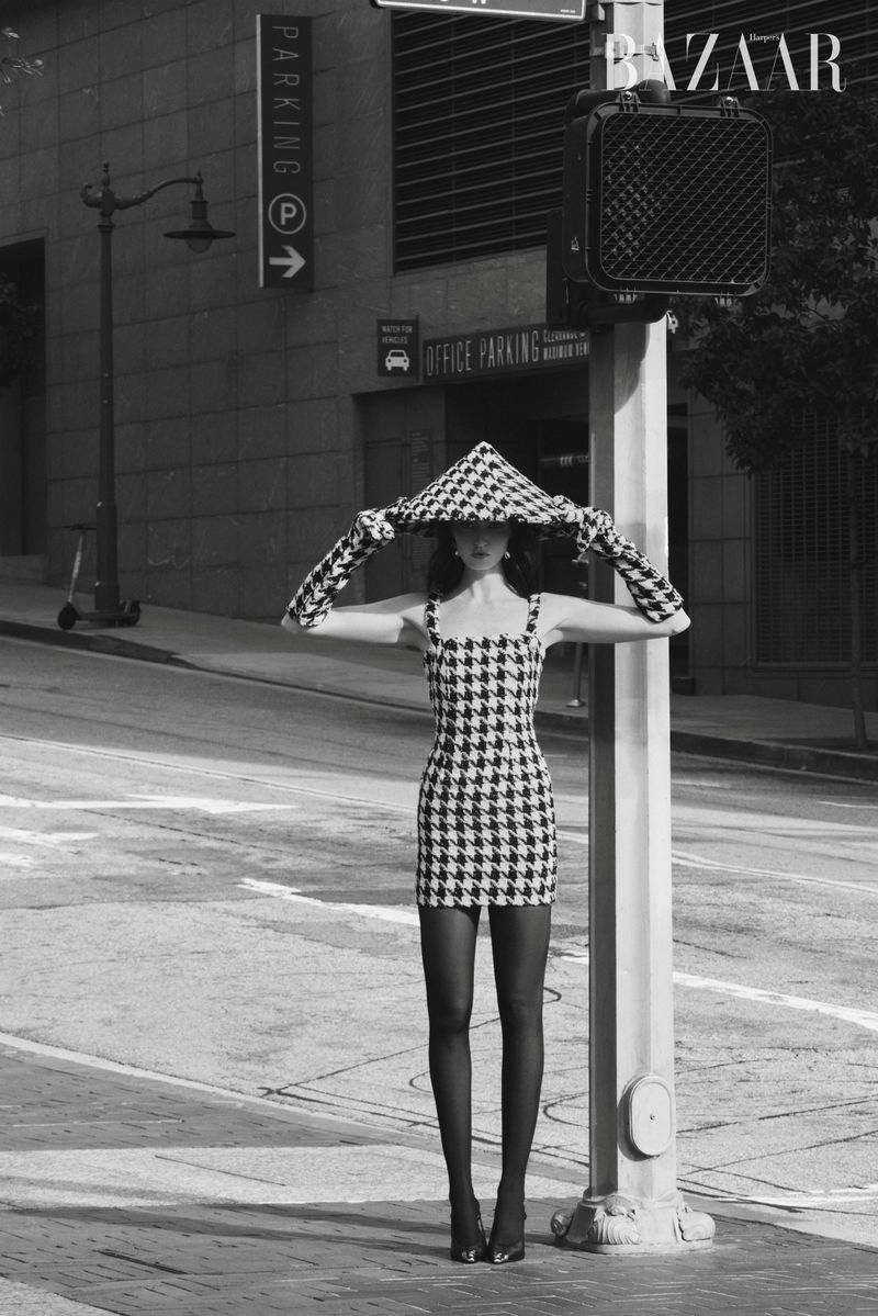 A black and white full-length shot of a woman standing on a street corner, wearing a houndstooth dress and hat with matching gloves.