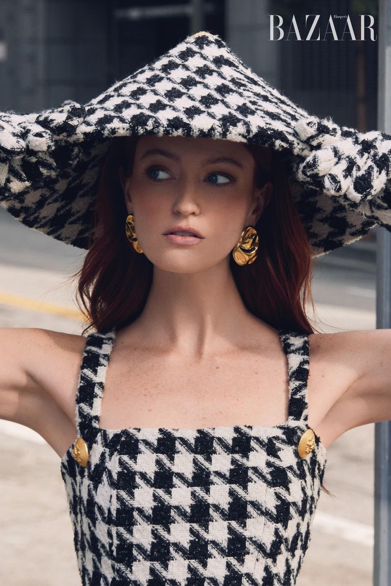 Fashionable woman poses in houndstooth hat and dress, accessorized with gold earrings, in an outdoor urban setting with muted colors.