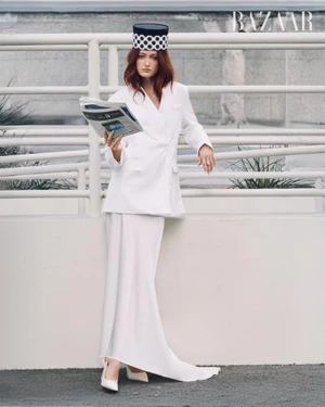 Model Megan Puleri in an all-white tailored suit and polka dot pillbox hat holding a newspaper by an outdoor railing.