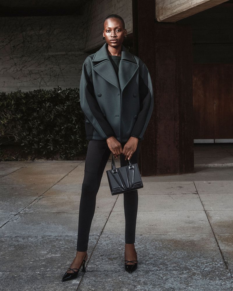 An adult female fashion model poses in an outdoor setting, wearing a stylish jacket, leggings, and heels, and carrying a sleek handbag.