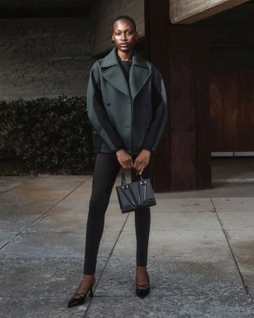 An adult female fashion model poses in an outdoor setting, wearing a stylish jacket, leggings, and heels, and carrying a sleek handbag.