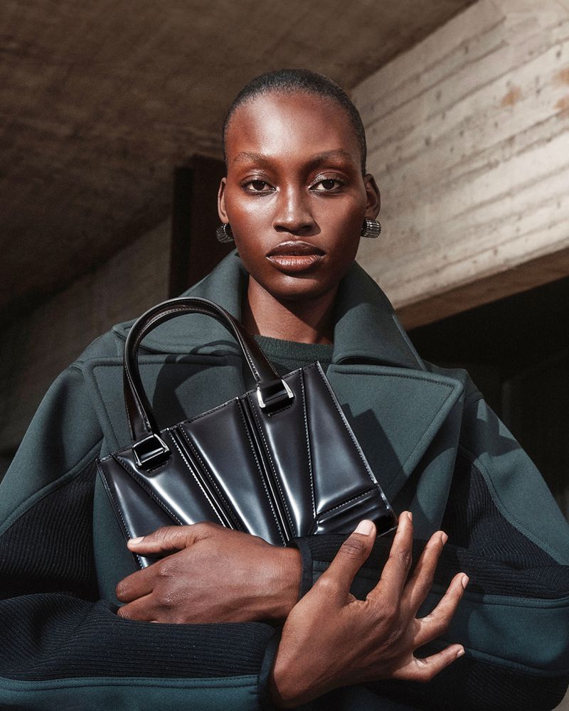 A striking portrait of a Black woman in a dark green coat holding a glossy black purse against an abstract architectural background.