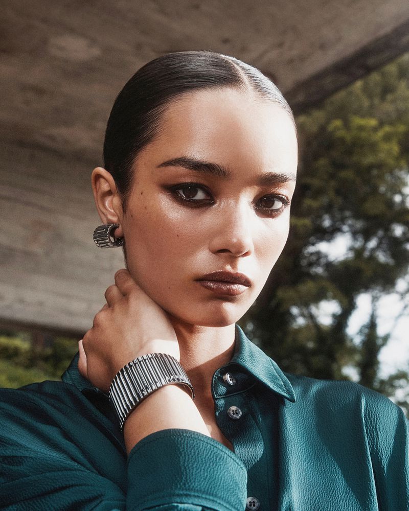 An adult woman wearing statement jewelry and a green collared shirt poses outdoors with a neutral expression, showcasing fashion and beauty.