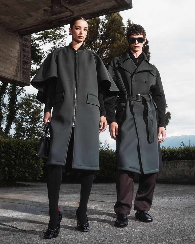 Two models pose outdoors wearing stylish green outerwear. Woman wears long-sleeved coat and heels, man wears sunglasses and belted coat.