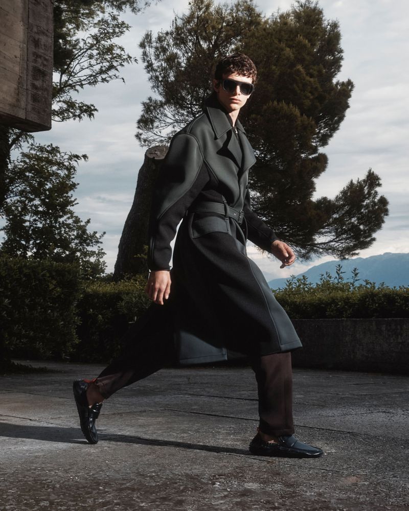 A stylish man in a long dark coat strides confidently on a concrete surface, with trees and a building in the background under a cloudy sky.