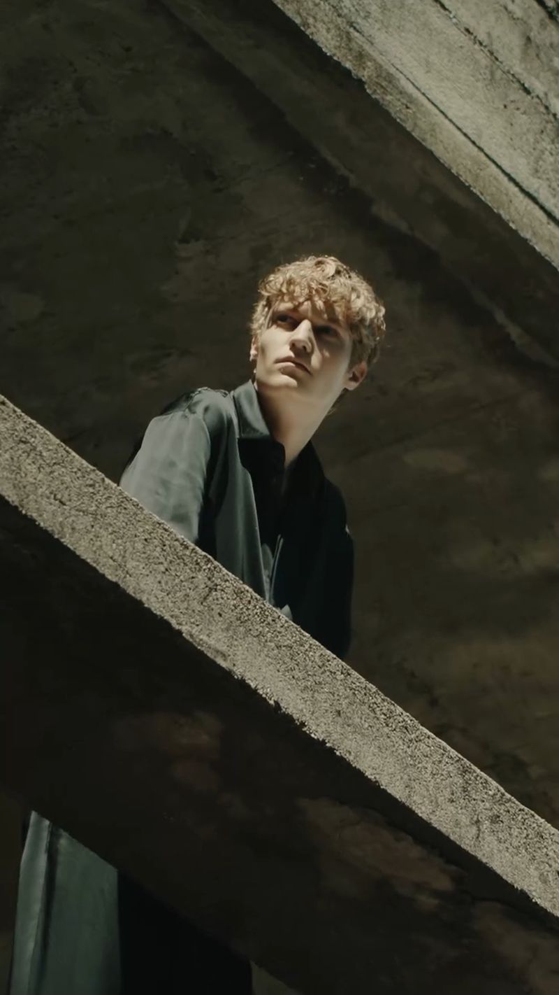 A young male model posing on a concrete structure in a dark architectural setting for Ferrari FW24 fashion campaign.