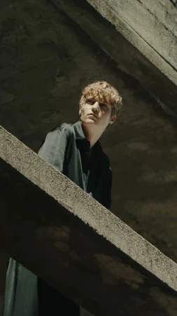 A young male model posing on a concrete structure in a dark architectural setting for Ferrari FW24 fashion campaign.