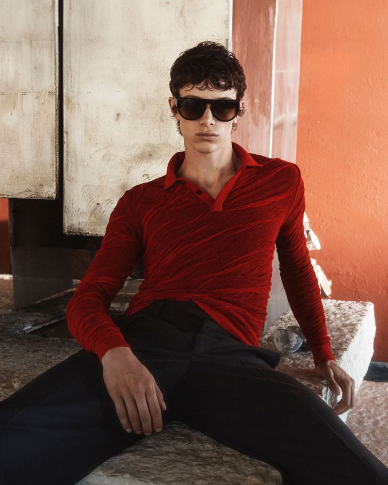 A young adult man is posing outdoors wearing sunglasses, a red shirt, and black pants, sitting on concrete blocks in an urban area.