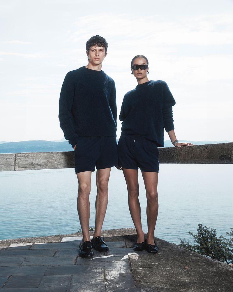 Two models in matching navy sweaters and shorts pose outdoors overlooking a tranquil ocean under a bright sky on a stone platform.