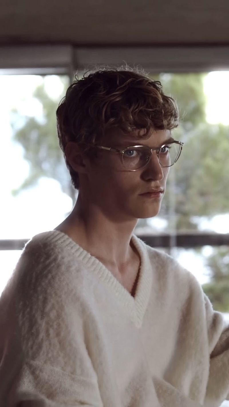 Young man wearing wire-rimmed glasses and a white V-neck sweater inside a room with large windows