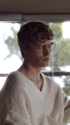 Young man wearing wire-rimmed glasses and a white V-neck sweater inside a room with large windows