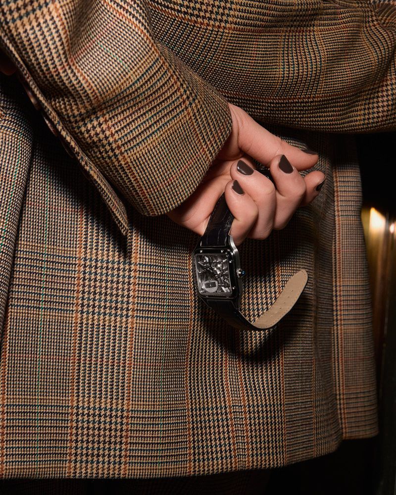 Close-up of a person in a plaid blazer holding a black wristwatch behind their back, showcasing fashion and accessory details.