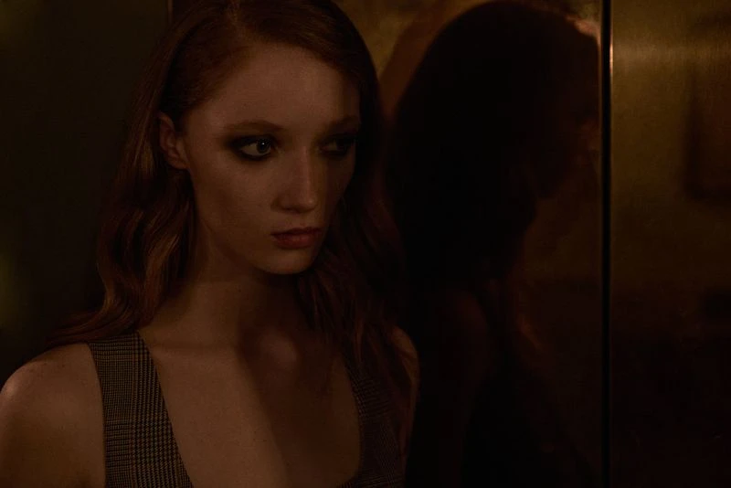 Portrait of a young redhead woman with smoky eyes and sleeveless top in a dimly lit indoor setting with reflective surface.