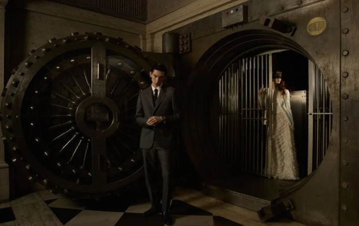 A well-dressed man stands near a large vault door, while a woman in a gown stands inside the open vault, creating a mysterious scene.