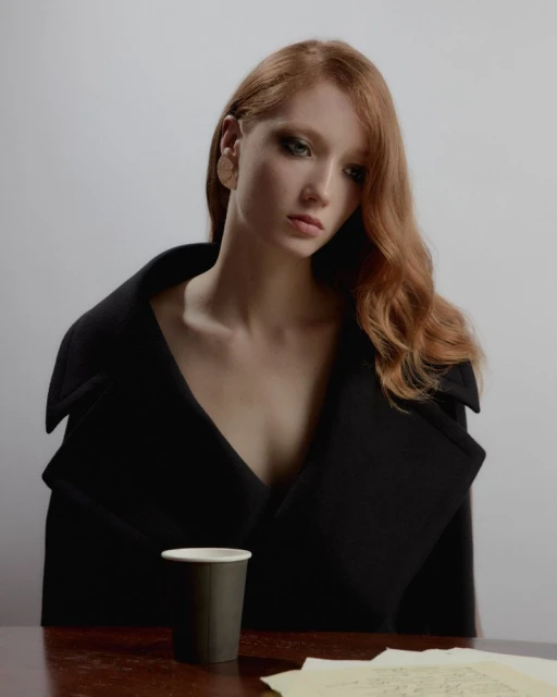 Portrait of a young woman with red hair wearing a black coat, seated at a table with a coffee cup and papers in a neutral setting.