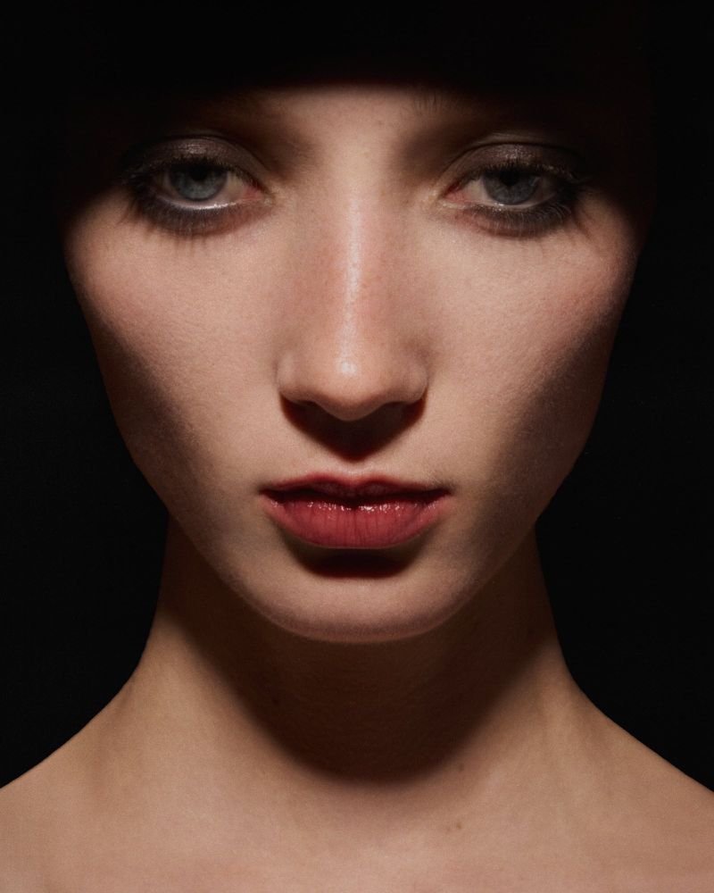 Dramatic portrait of a fair-skinned woman with blue eyes and dark lipstick, illuminated by a spotlight against a dark background.