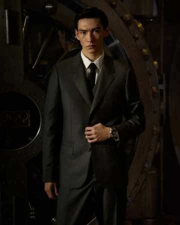 An Asian man in a dark suit stands in front of a large metal vault door, looking at the camera with a serious expression.