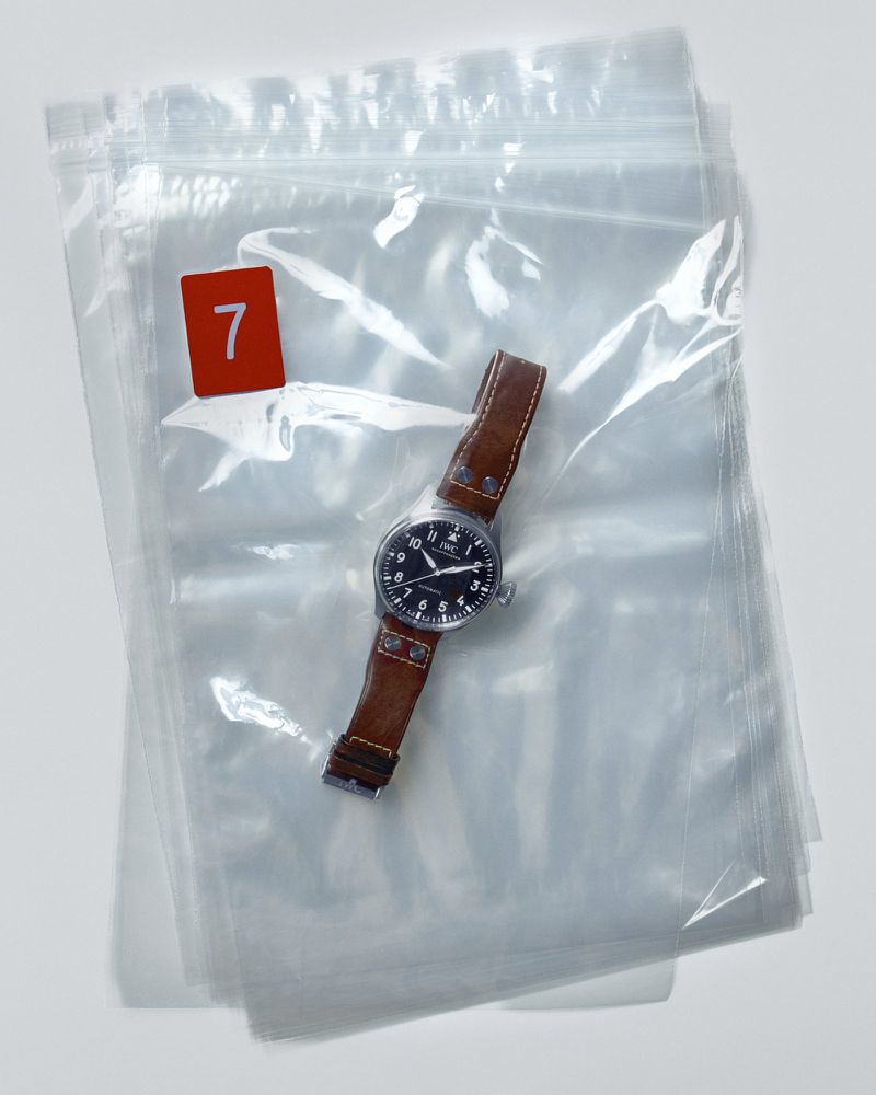 Analog wrist watch with a brown leather strap sits inside a clear plastic bag with a red number 7 visible on the top left.