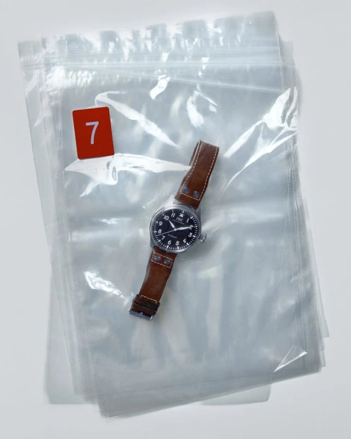 Analog wrist watch with a brown leather strap sits inside a clear plastic bag with a red number 7 visible on the top left.