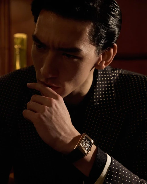 Close-up portrait of an adult male wearing a dotted jacket and a black wristwatch with a gold frame in dim, warm lighting.